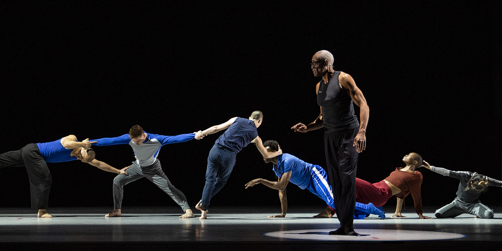 Six dancers in the background in various poses. One man in the foreground.