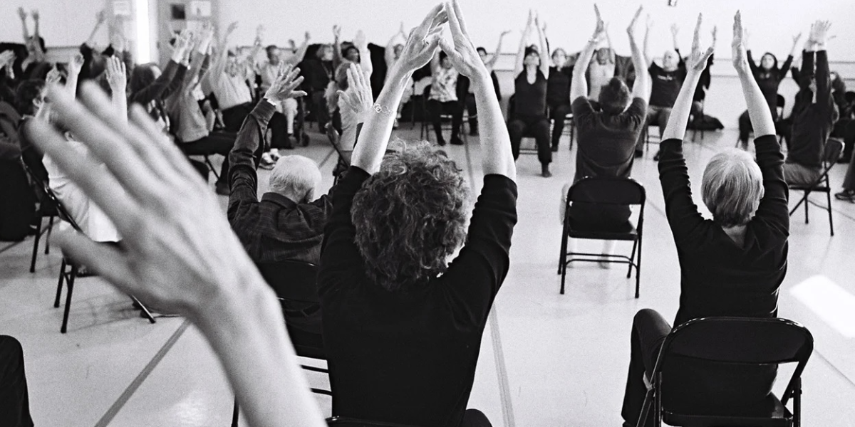 A black and white photo of people from the back sitting in chairs with their hands in the air.