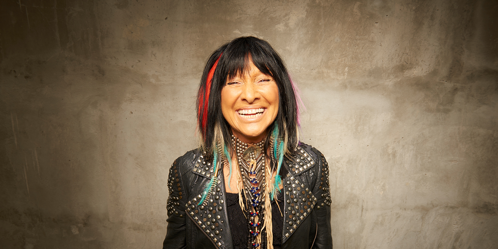 Buffy Sainte-Marie (a smiling woman with dark hair and leather jacket against a tan background)