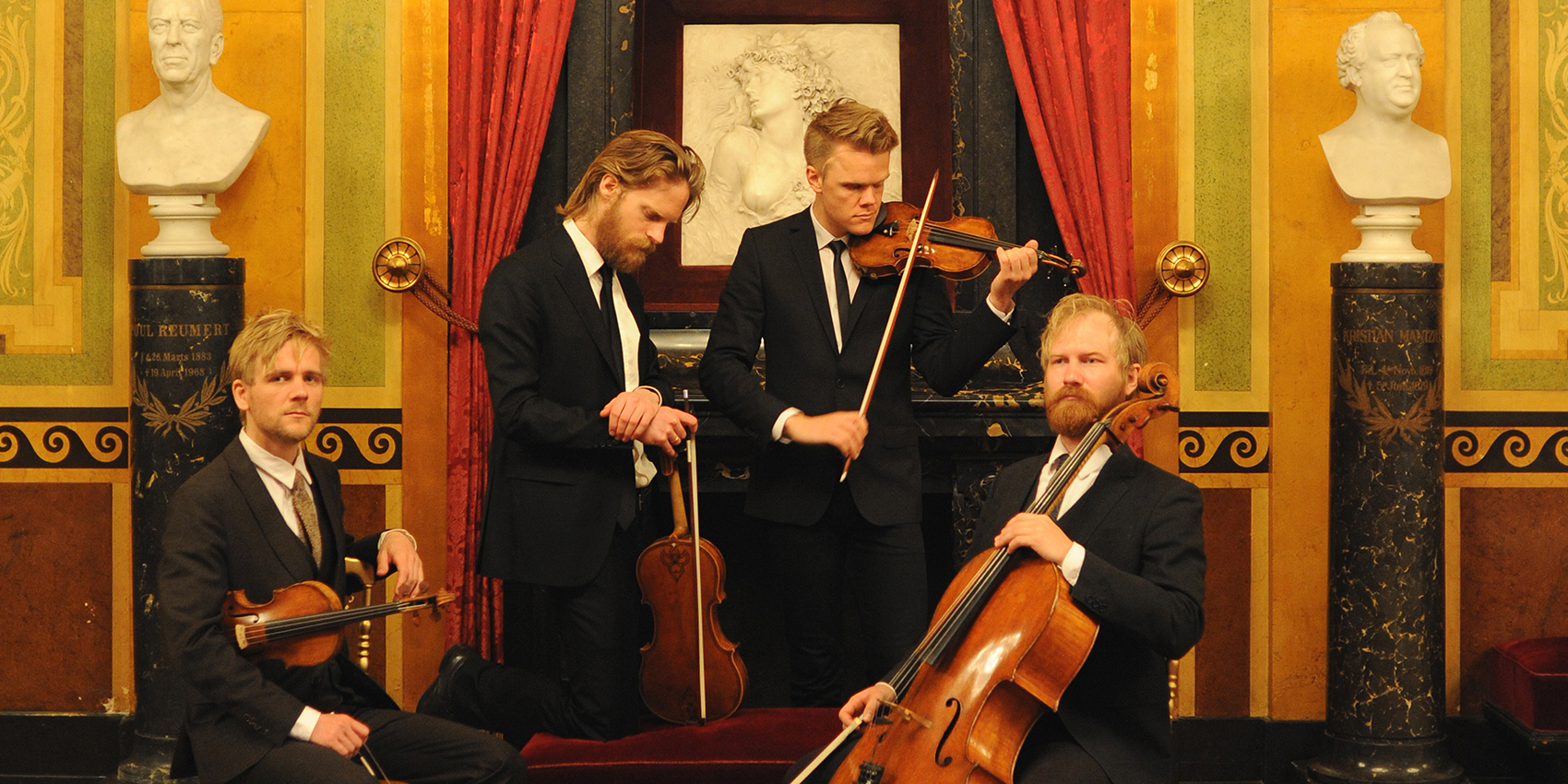 Four musicians holding string instruments in front of an ornate background