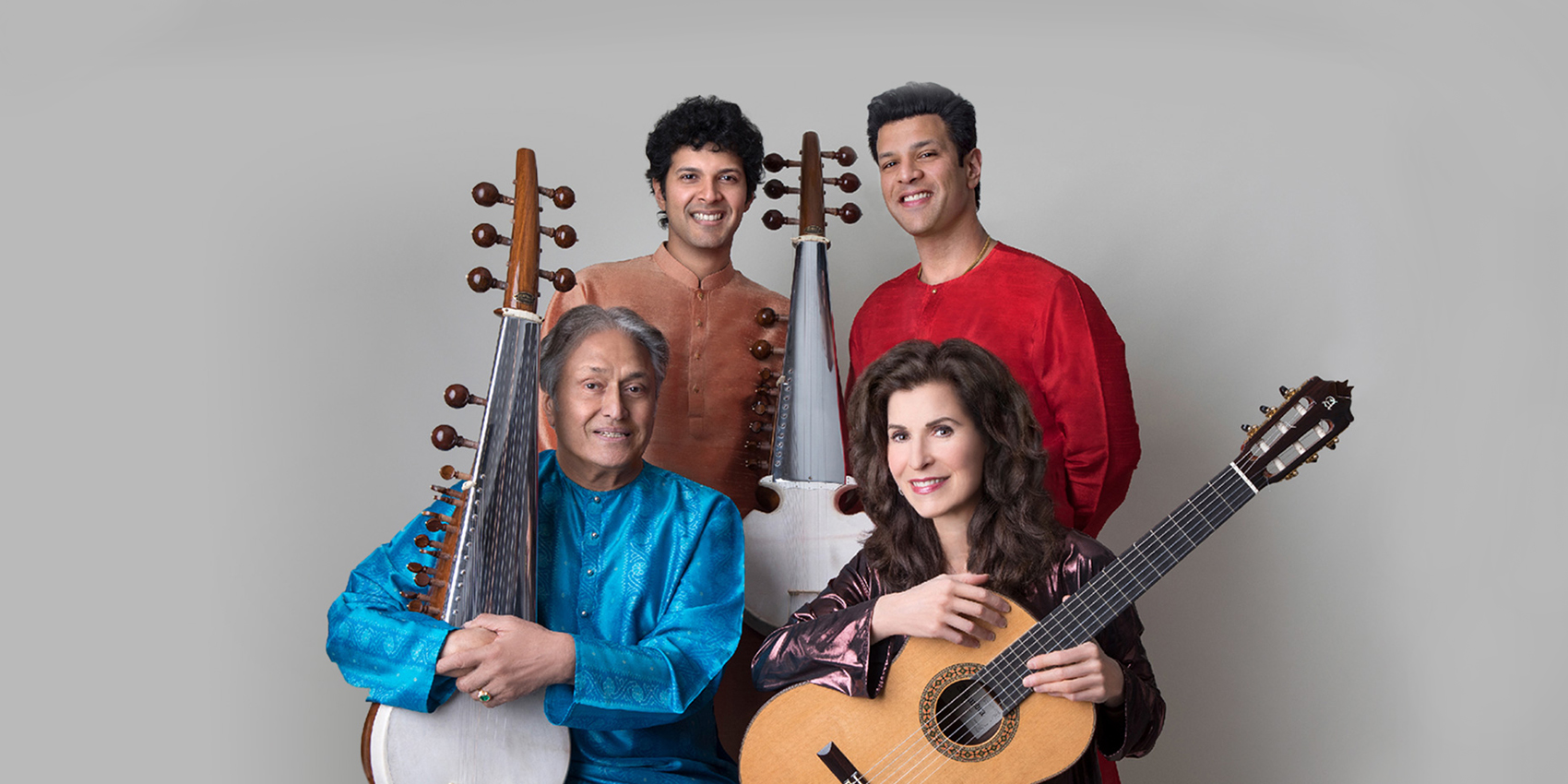 Four musicians, two sitting and two standing, holding instruments in front of a plain, gray background. 
