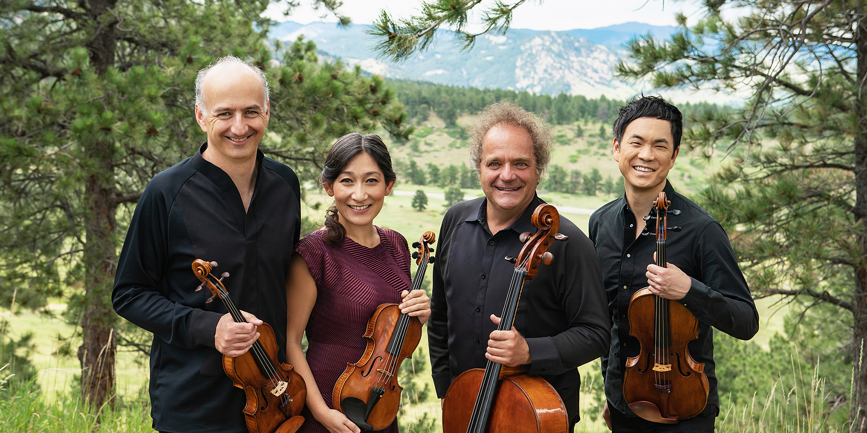 Four musicians standing, holding instruments outside. Trees and mountains in the background