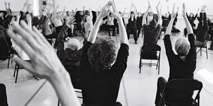 A black and white photo of people from the back sitting in chairs with their hands in the air.