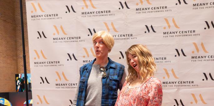 Two people dress as snl characters and pose in front of white background with logos
