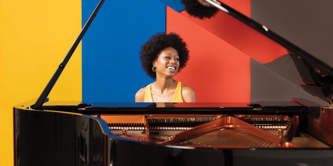 Isata Kanneh-Mason sitting at a grand piano against a colorful background