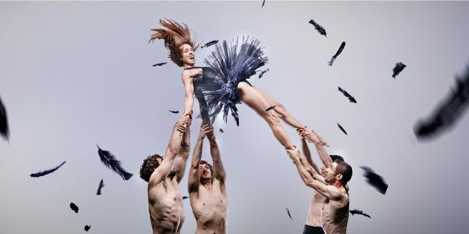 Four shirtless dancers lift dancer in black tutu, looking distressed. Feathers fly around. 