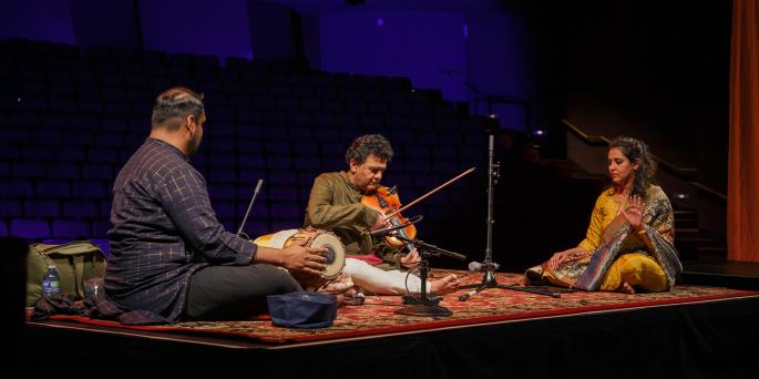 Violinist sits on rug and plays while two other instrumentalists sit beside