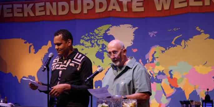 Two people stand in front of weekend update background alluding to Saturday Night Live