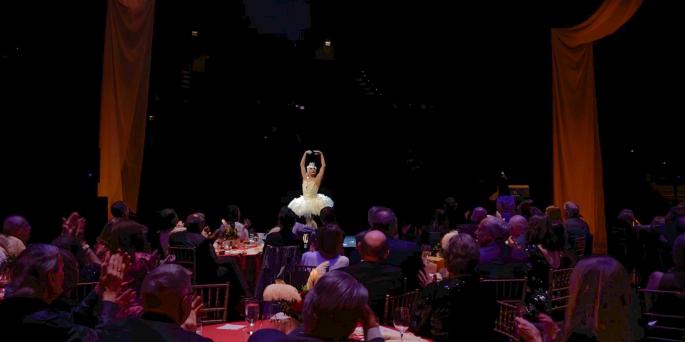 Ballerina in white raises arms in front of seated diners