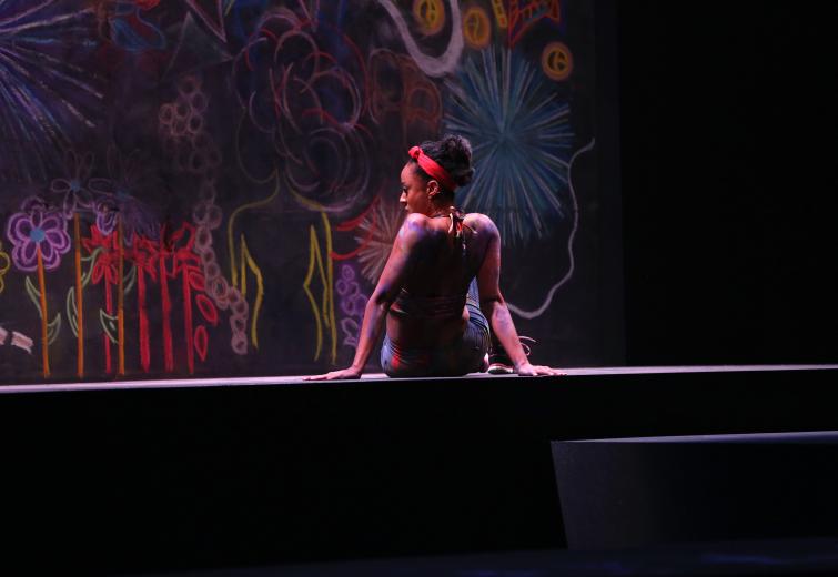 One female performer sits on stage in front of a colorful chalk wall