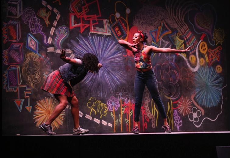 Two female dancers on stage in front of a colorful chalk wall