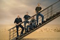 Four musicians stand in a desert scene on an industrial staircase