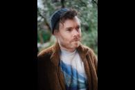 Gabriel Kahane in a sweater and beanie looking to the side of the camera